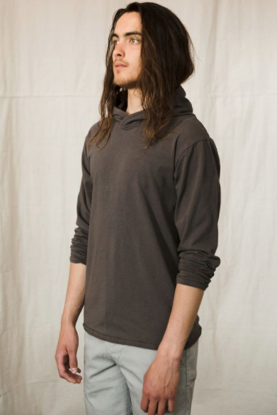 A handsome young man with long hair wears a charcoal grey hooded long sleeve pullover top with the sleeves pushd up